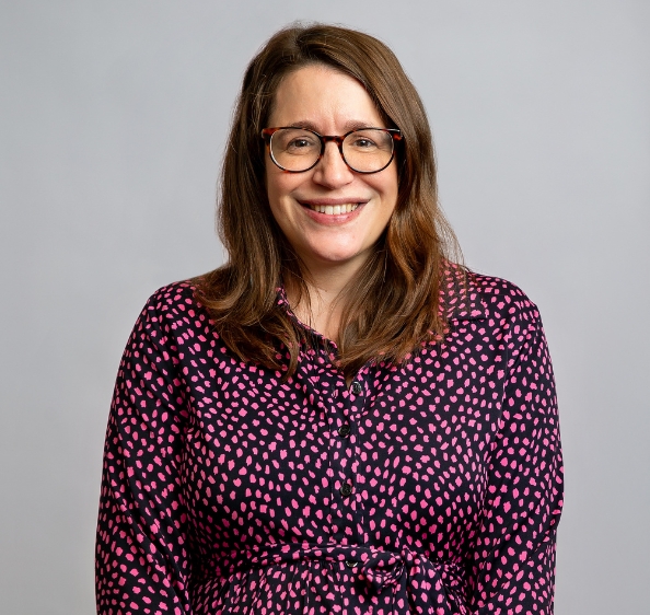 Female with shoulder length brown hair, wearing glasses and smiling. In pink and black patterned dress.